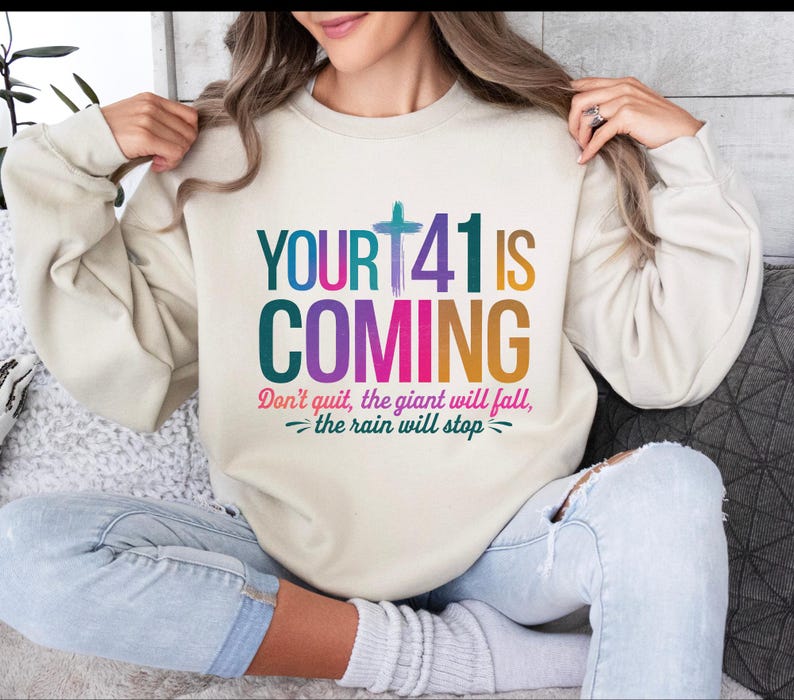 May include: A cream colored sweatshirt with the text "YOUR+41 IS COMING" in rainbow colors. The text "Don't quit, the giant will fall, -the rain will stop- " is printed below the main text.