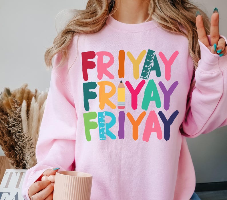 May include: Pink sweatshirt with the words "FRIYAY" repeated three times in rainbow colors. Each word has a different school supply graphic: a ruler, a pencil, and a ruler.