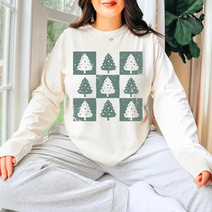 Puede incluir: Una camisa de manga larga de color crema con un patr&oacute;n de nueve &aacute;rboles de Navidad verdes con copos de nieve blancos. Los &aacute;rboles est&aacute;n dispuestos en una cuadr&iacute;cula de 3x3.
