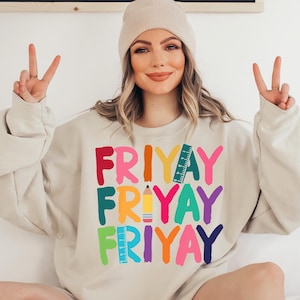 May include: A beige sweatshirt with the words "FRIYAY" repeated three times in colorful letters. Each word is decorated with a different school supply, including a pencil and a ruler.