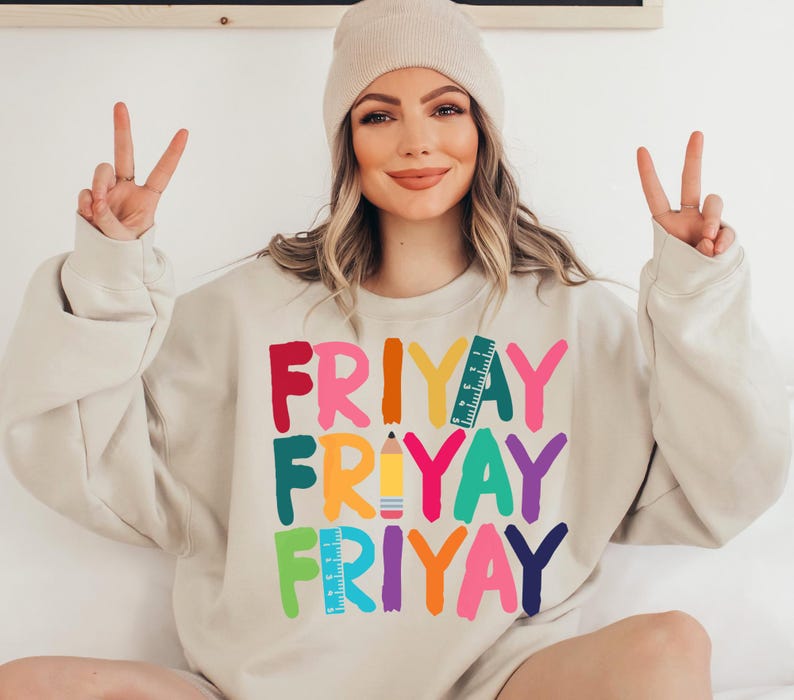 May include: A beige sweatshirt with the words "FRIYAY" repeated three times in colorful letters. Each word is decorated with a different school supply, including a pencil and a ruler.