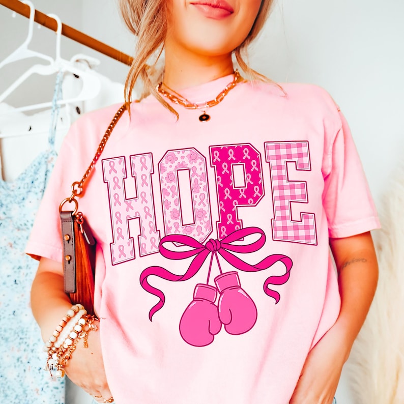Puede incluir: Camiseta rosa claro con la palabra "HOPE" en fuente decorativa. Las letras est&aacute;n adornadas con lazos rosas, motivos florales y un dise&ntilde;o de cuadros rosas. Debajo, unos guantes de boxeo rosas atados con una cinta rosa.