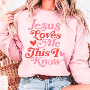 Puede incluir: Sudadera rosa con el texto "Jesus Loves Me This I Know" en una fuente retro con corazones rojos.