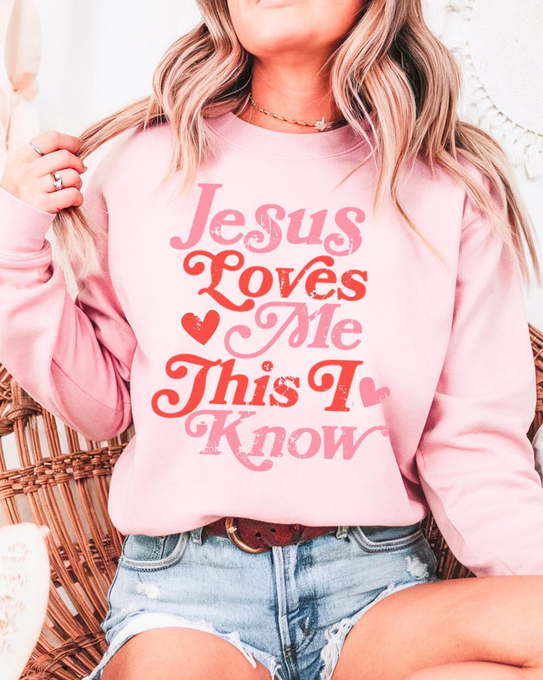 Puede incluir: Sudadera rosa con el texto "Jesus Loves Me This I Know" en una fuente retro con corazones rojos.