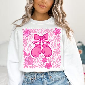 May include: White sweatshirt with a pink graphic featuring boxing gloves, a bow, flowers, and ribbons. The design is framed by pink floral and ribbon elements, creating a cohesive and supportive theme.