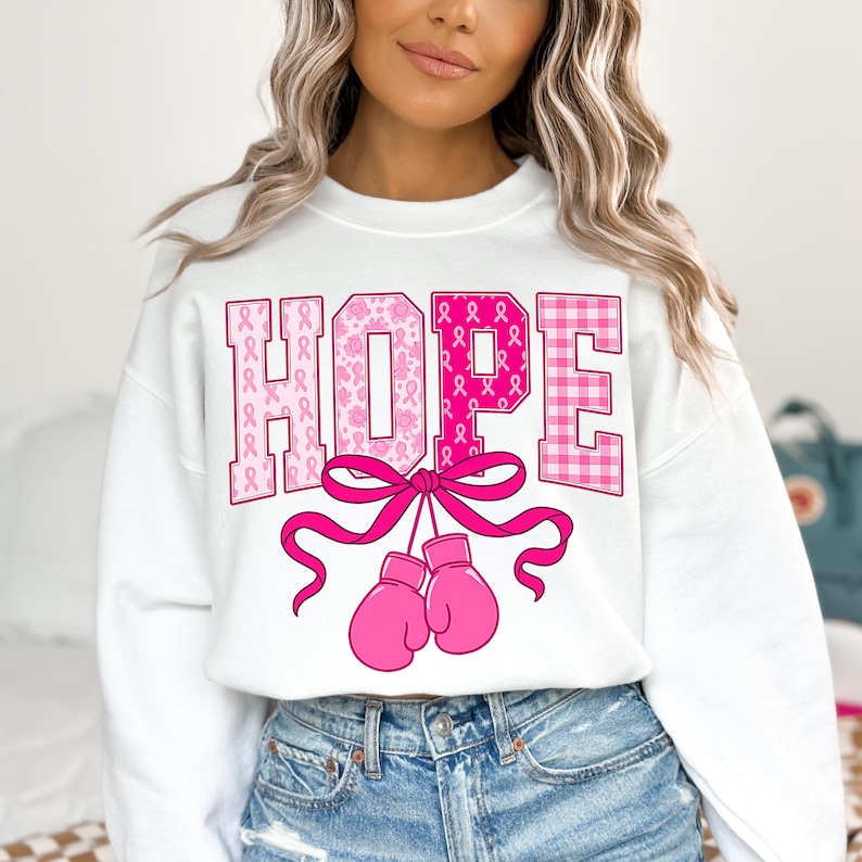 Puede incluir: Sudadera blanca con la palabra "HOPE" en rosa, con una mezcla de patrones que incluyen cintas y cuadros. Debajo de la palabra "HOPE" hay un lazo rosa y un par de guantes de boxeo rosas. La sudadera es de estilo casual.
