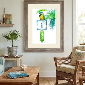 Coastal room decor mockup featuring a framed tropical watercolor print by David K Jones titled A Parrot Looks at 40. The artwork hangs in a room with a wicker chair and a Margaritaville planter with a small palm.