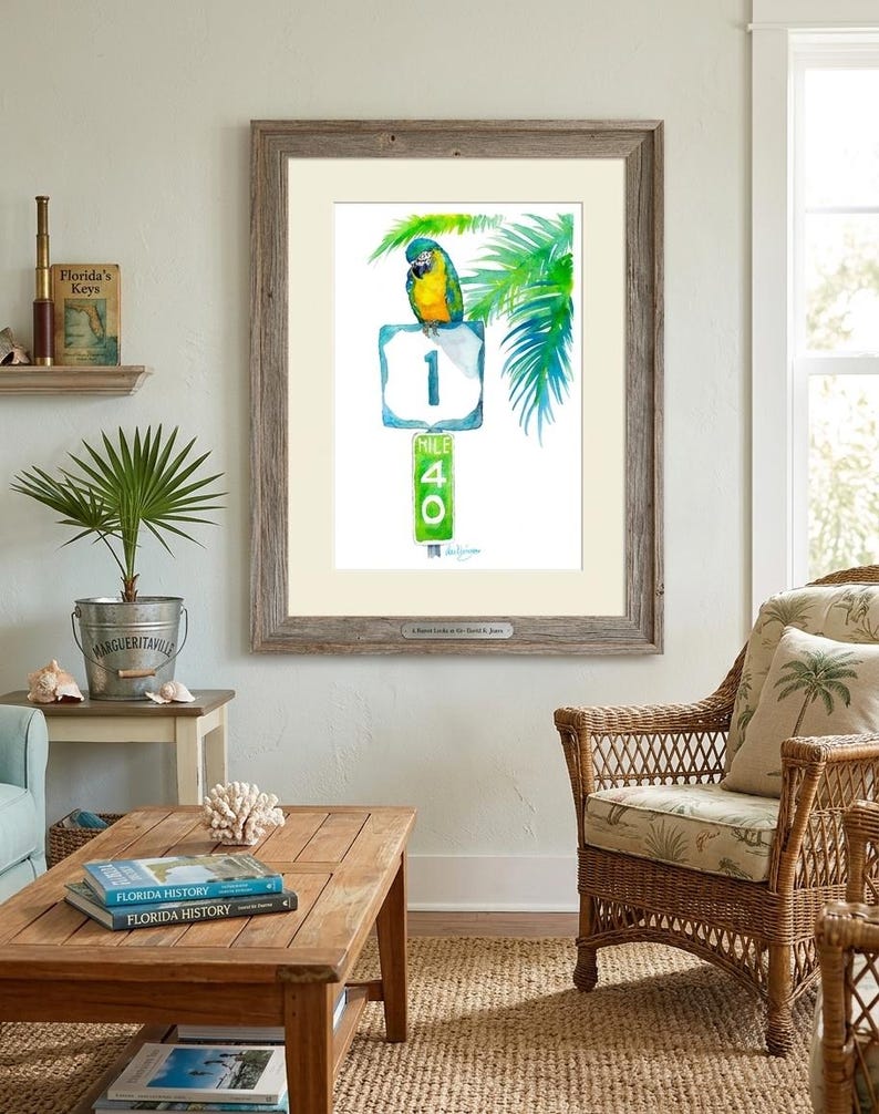 Coastal room decor mockup featuring a framed tropical watercolor print by David K Jones titled A Parrot Looks at 40. The artwork hangs in a room with a wicker chair and a Margaritaville planter with a small palm.
