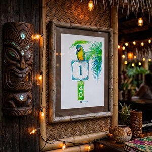 A framed Jimmy Buffett inspired watercolor art print, A Parrot Looks at 40 by David K Jones. A blue and yellow macaw sits on a US 1 highway shield above a green Mile Marker 40 sign, surrounded by tropical palm leaves in a coastal Florida Keys style. It is hung in a tiki bar wit thatched wall and a string of lights.