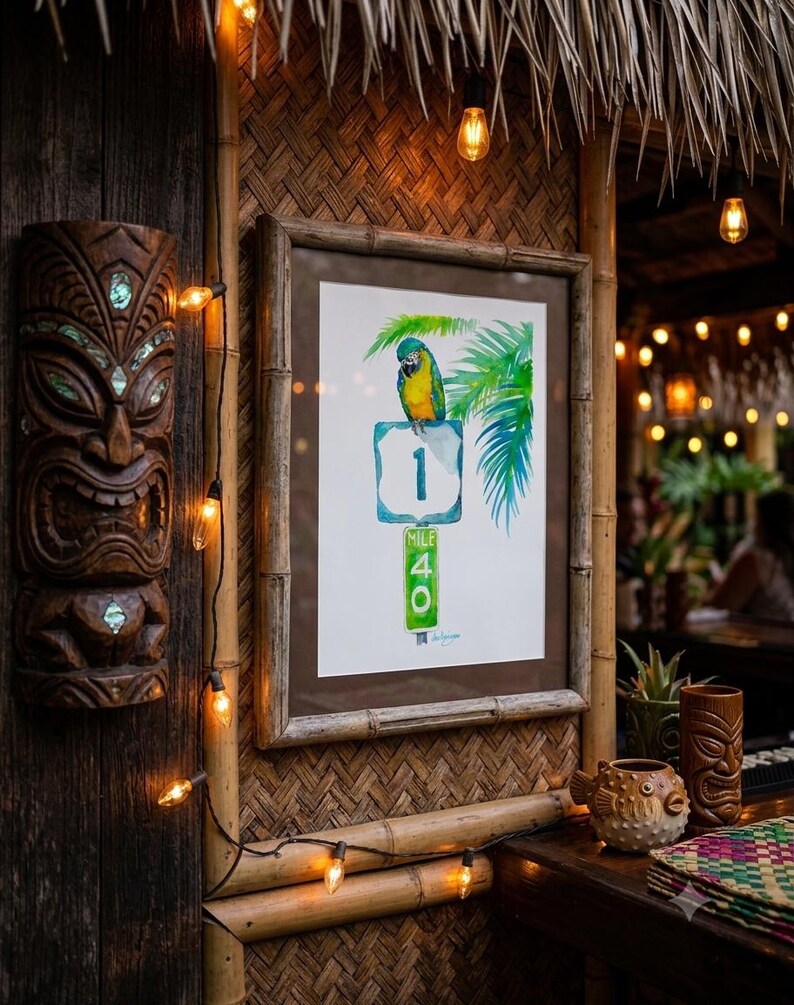A framed Jimmy Buffett inspired watercolor art print, A Parrot Looks at 40 by David K Jones. A blue and yellow macaw sits on a US 1 highway shield above a green Mile Marker 40 sign, surrounded by tropical palm leaves in a coastal Florida Keys style. It is hung in a tiki bar wit thatched wall and a string of lights.