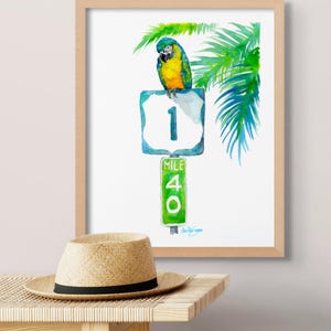 A wooden frame around a colorful watercolor of a parrot and palm tree on the Florida Keys mile maker 40 next to a table with a Panama hat by David K Jones.