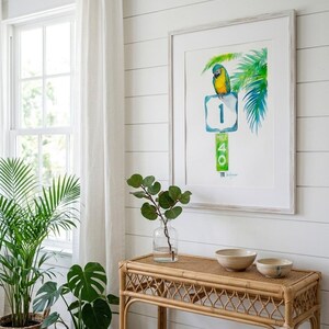 A framed Jimmy Buffett inspired watercolor art print, A Parrot Looks at 40 by David K Jones. A blue and yellow macaw sits on a US 1 highway shield above a green Mile Marker 40 sign, surrounded by tropical palm leaves in a coastal Florida Keys style. It is hung on a white wall in a bright coastal style room filled with tropical plants.