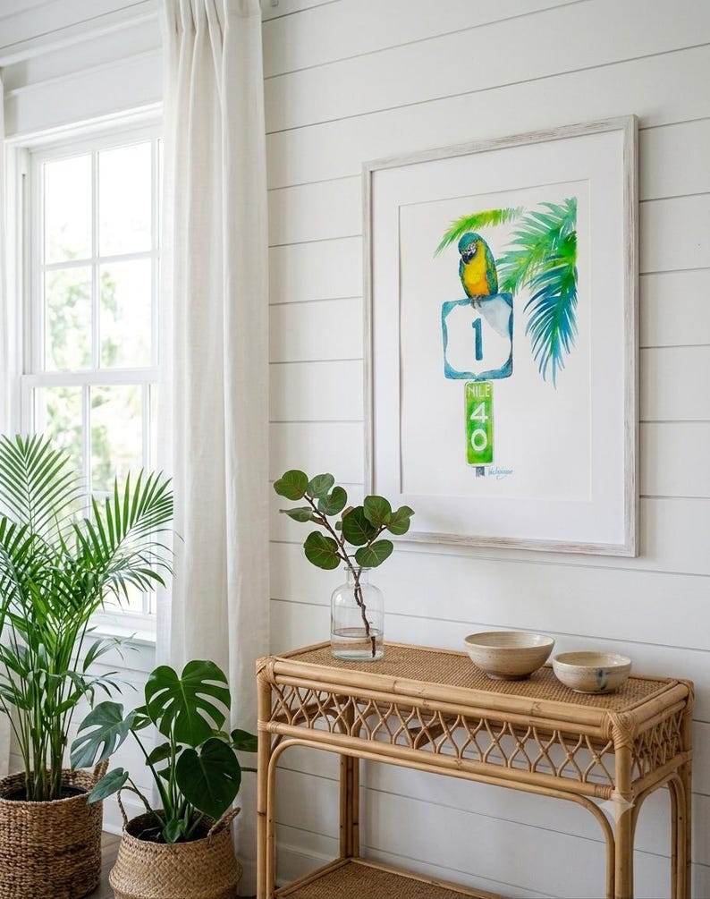 A framed Jimmy Buffett inspired watercolor art print, A Parrot Looks at 40 by David K Jones. A blue and yellow macaw sits on a US 1 highway shield above a green Mile Marker 40 sign, surrounded by tropical palm leaves in a coastal Florida Keys style. It is hung on a white wall in a bright coastal style room filled with tropical plants.