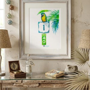 Coastal living room decor mockup featuring a framed tropical watercolor print by David K Jones titled A Parrot Looks at 40. The artwork hangs above a rustic wood entryway table decorated with a Panama hat, glass fishing floats, and a decorative bottle labeled Key West sand, creating a beach house aesthetic.