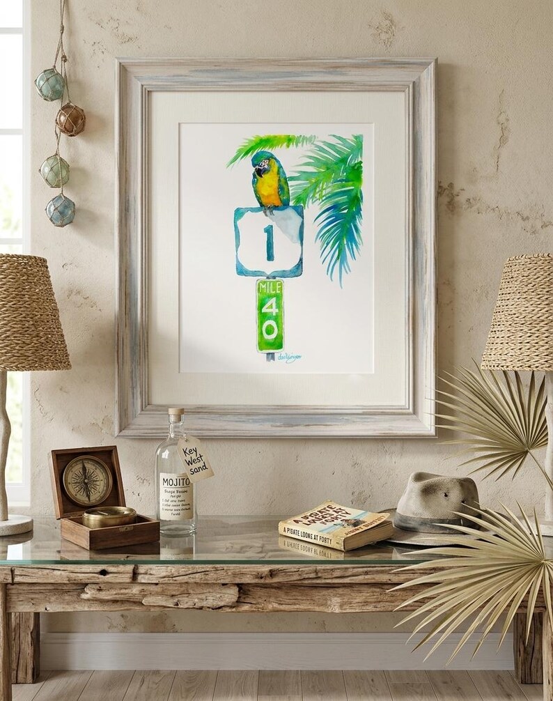 Coastal living room decor mockup featuring a framed tropical watercolor print by David K Jones titled A Parrot Looks at 40. The artwork hangs above a rustic wood entryway table decorated with a Panama hat, glass fishing floats, and a decorative bottle labeled Key West sand, creating a beach house aesthetic.