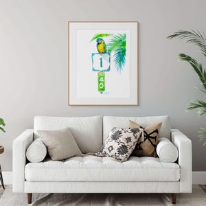 Florida-style living room decor mockup featuring a framed tropical watercolor print by David K Jones titled A Parrot Looks at 40. The artwork hangs above a comfortable white sofa with nautical themed cushions and tropical plants on either side giving a coastal decor feel.