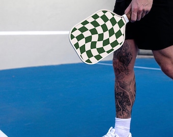 Könnte beinhalten: Eine Person hält einen weißen und grünen karierten Paddleball-Schläger auf einem blauen Tennisplatz.