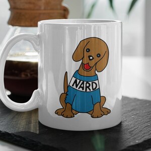 May include: White ceramic mug with a cartoon illustration of a brown dog wearing a blue shirt with the word "NARD" printed on it.
