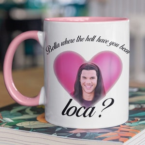 Puede incluir: Taza de cerámica blanca con asa y borde rosa. La taza presenta un corazón rosa con la cara de un hombre dentro. El texto en la taza dice "Bella where the hell have you been loca?"