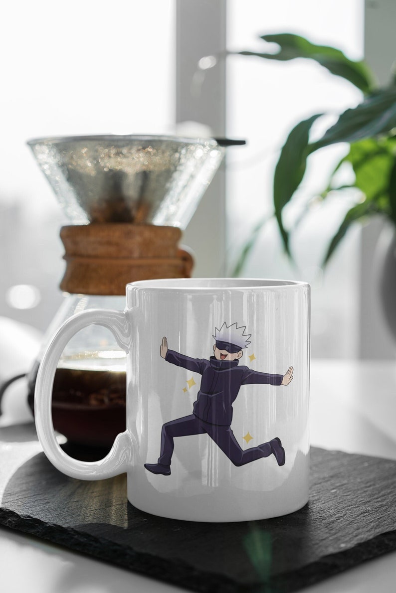 May include: White ceramic mug with a cartoon illustration of a character with white hair and a black eye mask. The character is wearing a black jumpsuit and is jumping with their arms outstretched. The mug has a black handle.