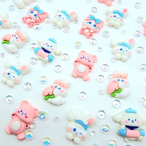 May include: A collection of small, colorful resin figurines, including bunnies, bears, and other cute characters. The figurines are primarily white and pink, with blue accents. Iridescent, clear beads are scattered around the figurines.