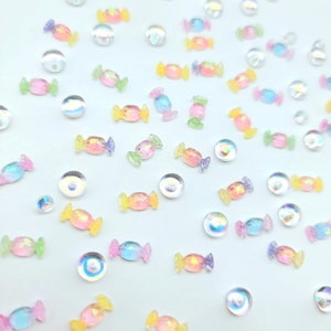 May include: Assortment of iridescent candy-shaped embellishments in various colors, including pink, blue, yellow, and green, scattered on a white background.