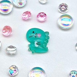 Può includere: Una statuetta di dinosauro verde acqua con una guancia rosa e occhi neri. La statuetta è circondata da perline di vetro iridescenti e trasparenti.