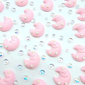 Puede incluir: Una colección de dijes en forma de media luna rosa con estrellas blancas y caras dormidas. Los dijes están esparcidos sobre un fondo blanco con brillantina iridiscente.