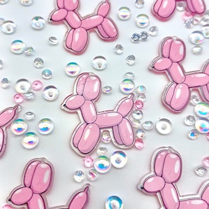 May include: Pink balloon animal charms in the shape of a dog. The charms are scattered on a white background with iridescent sequins.