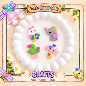 Puede incluir: Cuatro dijes de silicona coloridos que presentan una nube rosa con tentáculos de arcoíris, un elefante gris con orejas rosas, un cactus verde en una maceta rosa y un paraguas rosa con una nube. El texto Yua's Super Cute y CRAFTS es visible.