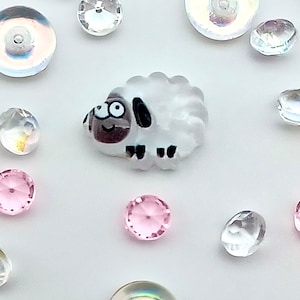 May include: A white ceramic sheep figurine with black eyes and a brown nose. The sheep is surrounded by iridescent and clear glass beads.