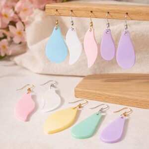 May include: A selection of pastel-coloured earrings in teardrop and leaf shapes. The earrings are displayed on a wooden stand. Colours include blue, white, pink, yellow, green, and purple. The earrings have gold and silver hooks.