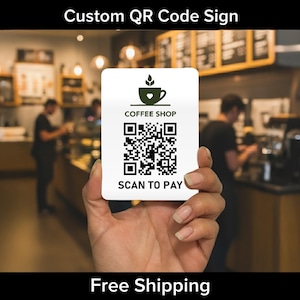 May include: A white rectangular sign with a QR code for a coffee shop, held in front of a blurred coffee shop background. The sign has the text "COFFEE SHOP" and "SCAN TO PAY". The top of the sign has a green coffee cup logo.