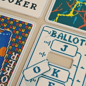May include: Close-up of playing cards with pixelated designs. The cards feature blue, yellow, red, and white graphics. One card reads "JOKER" in black pixelated letters. Another card has the word "BALLOT" in blue pixelated letters.