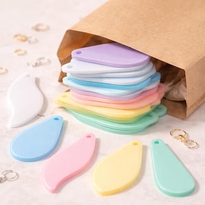 May include: A selection of pastel-coloured, teardrop-shaped keyrings. The keyrings are in shades of white, blue, pink, yellow, and green. A stack of keyrings is visible inside a brown paper bag. Several individual keyrings are scattered around the bag.