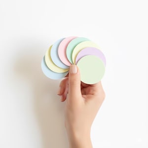 May include: A hand holds a fan of pastel-colored, circular paper cutouts against a white background. The colors include light blue, yellow, pink, green, and lavender. The paper circles are arranged in a semi-circular pattern.