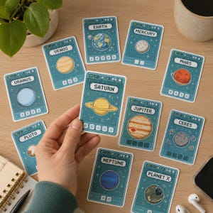 May include: A collection of teal-colored planet cards with pixelated illustrations of the planets. The cards feature names like "Saturn," "Earth," and "Venus." A hand holds one card, with other cards scattered on a wooden surface.