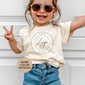 May include: A young child wearing a light beige t-shirt with a circle graphic that says "Your Logo Here" in black script. The child is wearing blue denim shorts and has a pink bow in their hair. The child is smiling and making peace signs with both hands. The background is a white wall.