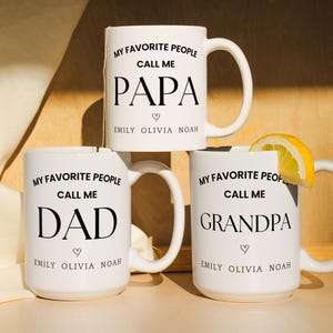 May include: Three white ceramic mugs stacked on top of each other. The mugs have black text that reads "MY FAVORITE PEOPLE CALL ME PAPA", "DAD", and "GRANDPA". Each mug has a heart symbol and a name below the text. A lemon slice rests on the top mug.