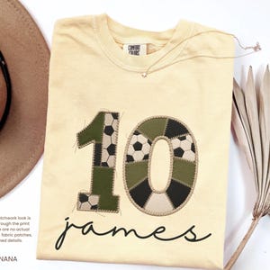 May include: A light yellow t-shirt featuring a large number 10 with a faux patchwork design of soccer ball patterns and olive green fabric. Below the number, the name James is printed in black cursive script. The shirt has a Comfort Colors label.