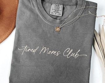 Può includere: Una maglietta grigia Comfort Colors con la scritta "Tired Moms Club" in corsivo. Una collana color oro con un ciondolo è appoggiata sulla maglietta. Un cappello marrone e steli di piante secche sono sullo sfondo.