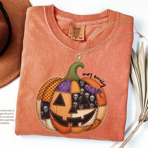May include: A burnt orange short-sleeve t-shirt featuring a large graphic of a faux patchwork pumpkin with a jack-o'-lantern face, skull patterns, and purple bat accents. The text "mrs emily" is printed in black script next to the pumpkin.