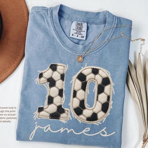 May include: A light blue crew neck t-shirt featuring a large graphic of the number 10 designed with a black and white soccer ball pattern. Below the number, the name James is printed in white script. The shirt has a Comfort Colors label.