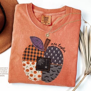 May include: A burnt orange t-shirt featuring a printed faux patchwork apple design. The apple graphic includes patterns of orange and black buffalo plaid, pumpkins, skulls, bats, and a spiderweb. The text 'mrs taylor' is printed in black script.