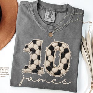 May include: A charcoal gray short-sleeve t-shirt featuring a large graphic of the number 10 designed with a black and white soccer ball pattern. Below the number, the name James is printed in a light beige script font. The shirt has a crew neckline.