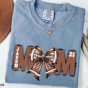 May include: Blue jean colored t-shirt with a football-themed design. The design includes the word "MOM" with a football bow in the center, the name "JASON" on the left, and the number "21" on the right. A gold necklace is draped across the shirt.