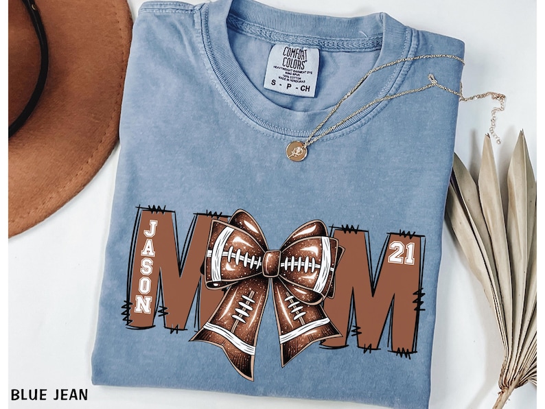 May include: Blue jean colored t-shirt with a football-themed design. The design includes the word "MOM" with a football bow in the center, the name "JASON" on the left, and the number "21" on the right. A gold necklace is draped across the shirt.