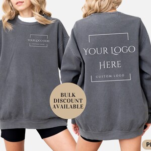 May include: A dark gray sweatshirt with the text "YOUR LOGO HERE" printed on the front and back. The front of the sweatshirt also has the text "CUSTOM LOGO". The sweatshirt is a Pepper color and is available in bulk.