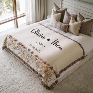 May include: A cream-colored personalized throw blanket with floral accents and fringed edges. The blanket features the names "Olivia & Theo" and the date "12.04.2025" in elegant script, perfect for a wedding or anniversary gift.