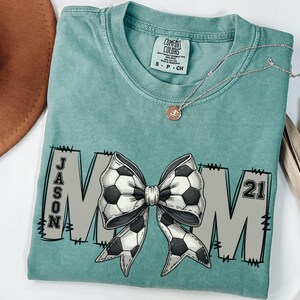 May include: Light green t-shirt with a large white and black bow with a soccer ball pattern. The bow is centered on the shirt with the letters "MOM" on either side. The left side of the shirt has the name "JASON" and the right side has the number "21".